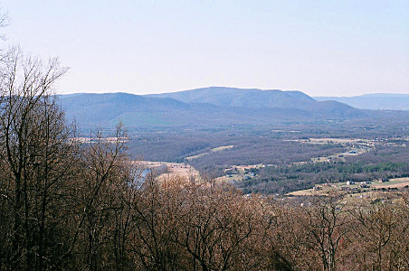 [There is a row of trees along the left and the lower edge of the image. Beyond that is a valley section with sections of trees and sections of what appears to be farm fields. In the distance are several levels of smooth-top mountains covered in trees. The further sections are less distinct due to haze.]